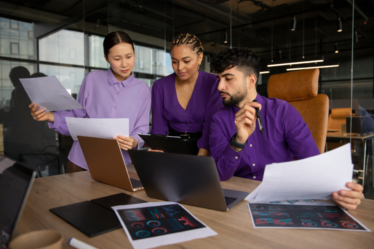 Três pessoas conversando ao redor de uma mesa de escritório com dois notebooks em cima, um homem sentado em frente a um notebook, duas mulheres em pé, uma com papeis nas mãos e a outra com um tablet em mãos, os três conversando sobre o que está nos papeis e no tablet em ambiente de escritório representando: Obrigações acessórias e responsabilidade solidária.