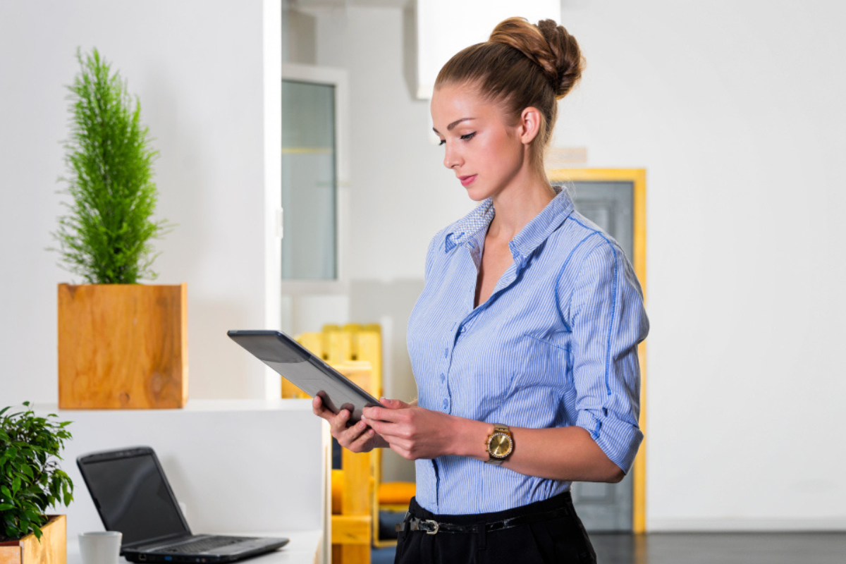 Mulher usando tablet em pé com uma mesa ao lado e um notebook sobre a mesma representando: Como evitar erros na emissão de notas fiscais durante a transição tributária.