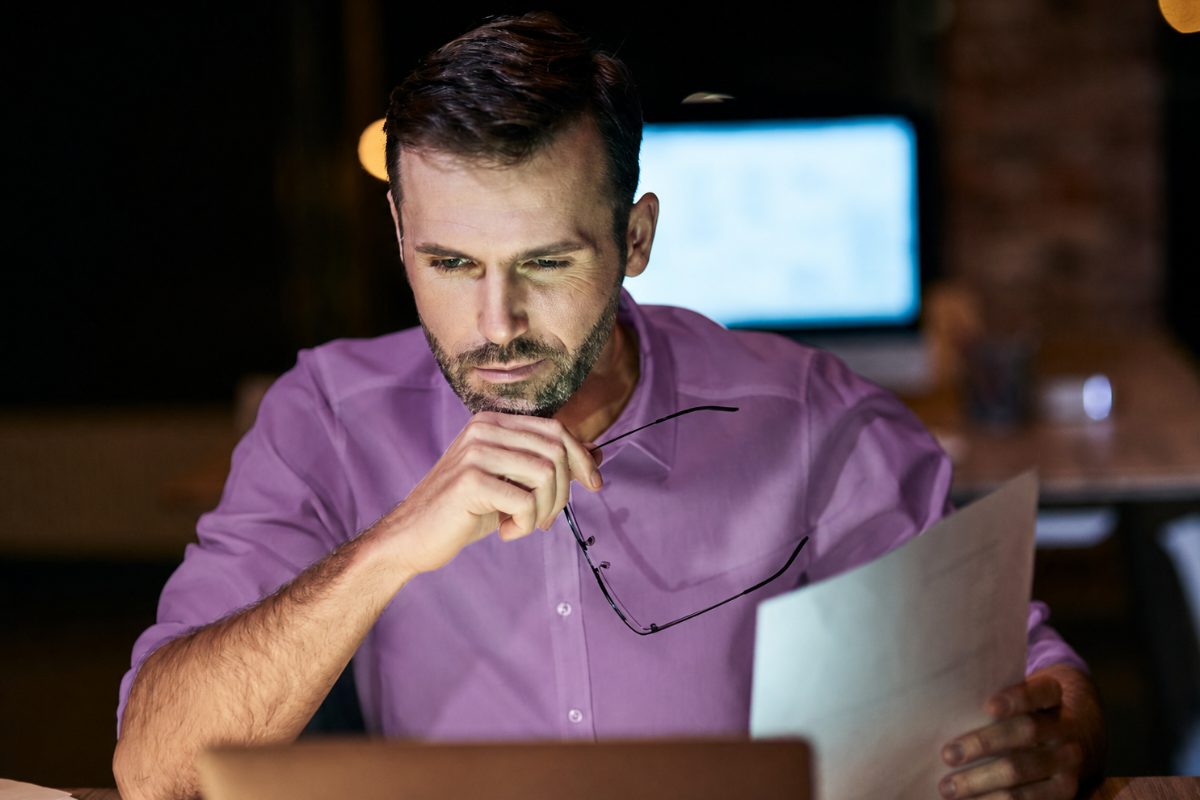 Homem trabalhando em ambiente de escritório em seu computador e analisando documentos em mãos e ao fundo outra mesa com um computador possivelmente do colega de trabalho, representando: O que avaliar antes de contratar um ERP?