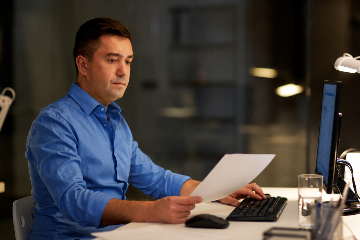 Empresário trabalhando em notebook e lendo documento em mãos em ambiente de escritório representando: Parametrização fiscal: por que isso é decisivo?