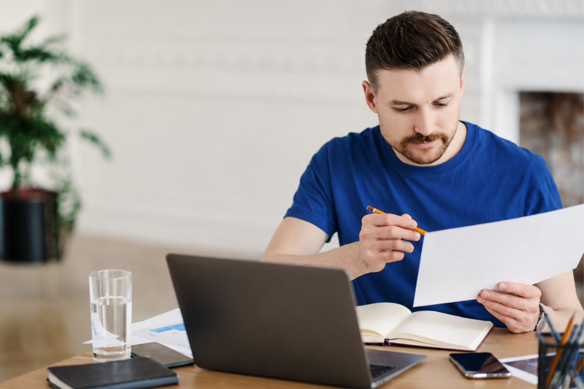 Homem trabalhando em seu computador e lendo documentos analisando Como formalizar seu negócio de revenda de software: MEI, ME ou LTDA?