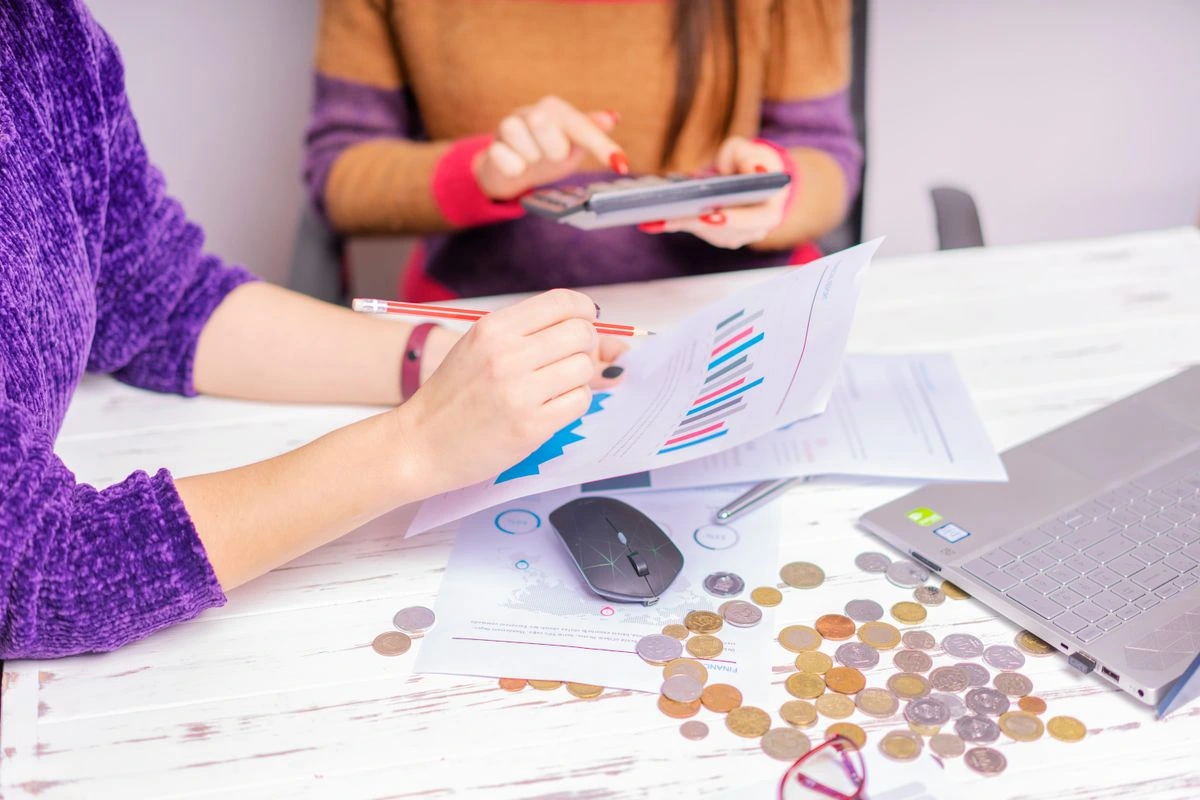 Duas mulheres usando um notebook, calculadora, caneta e analisando documentos e gráficos impressos em papeis representando: Precificação de honorários contábeis: como usar os dados do ERP dos clientes para cobrar pelo valor entregue.